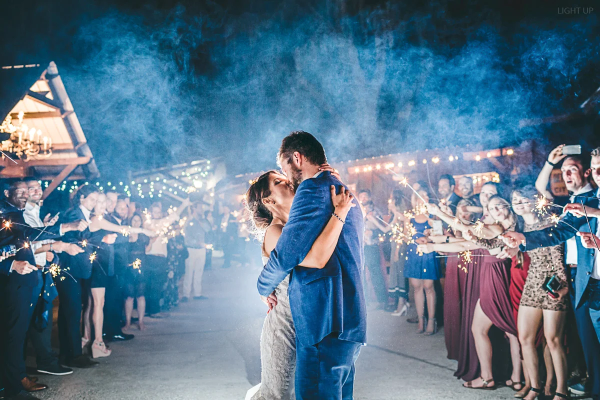 Wedding couple embraces with tight hug and kiss during while people around them wave sparklers for their sendoff at Club Lake venue