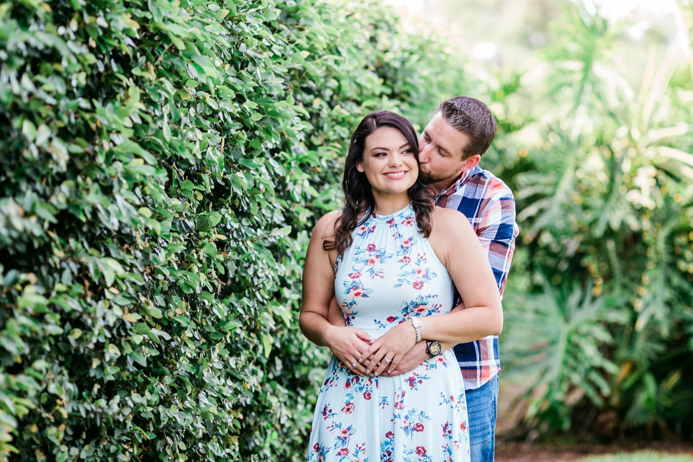 Man hugs and kisses his fiance from behind while standing in front of a green hedge at UCF Orlando for their engagement session