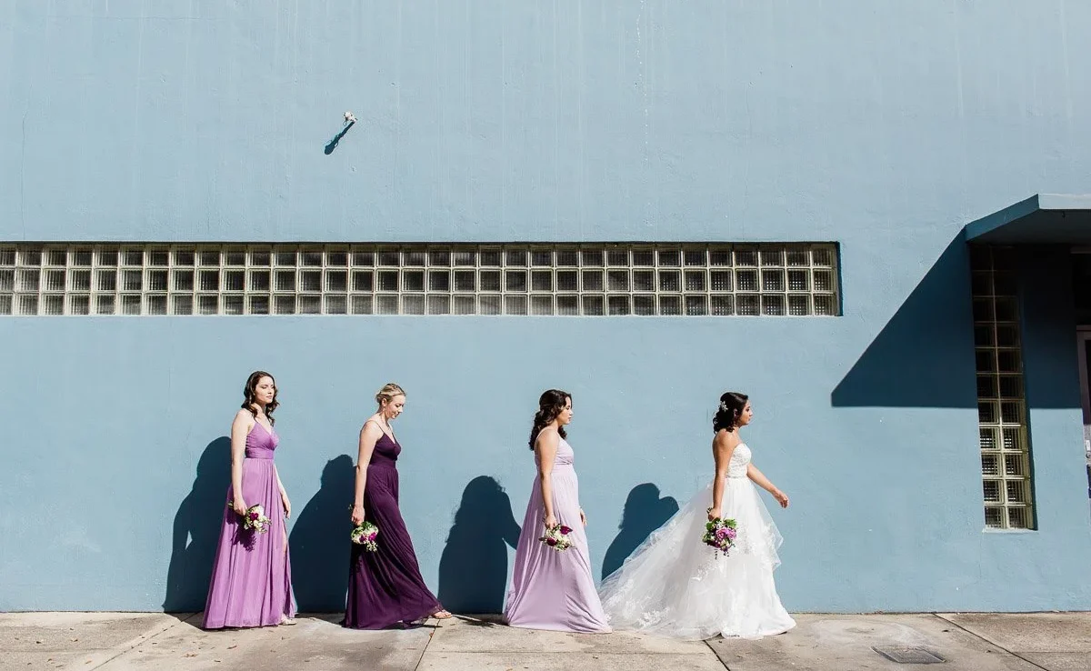 Bride and bridesmaids in purple color palate bridesmaids dresses walk on a sidewalk in front of a blue wall in downtown Winter Park FL
