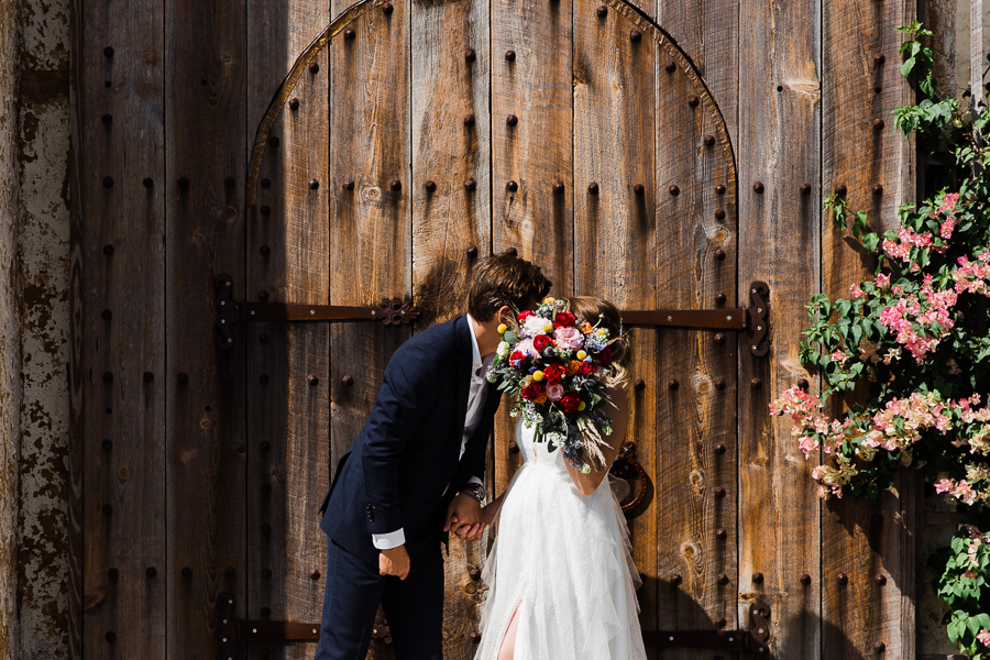Bride and groom kiss while hiding behind beautiful flower bouquet at Bella Cosa wedding venue