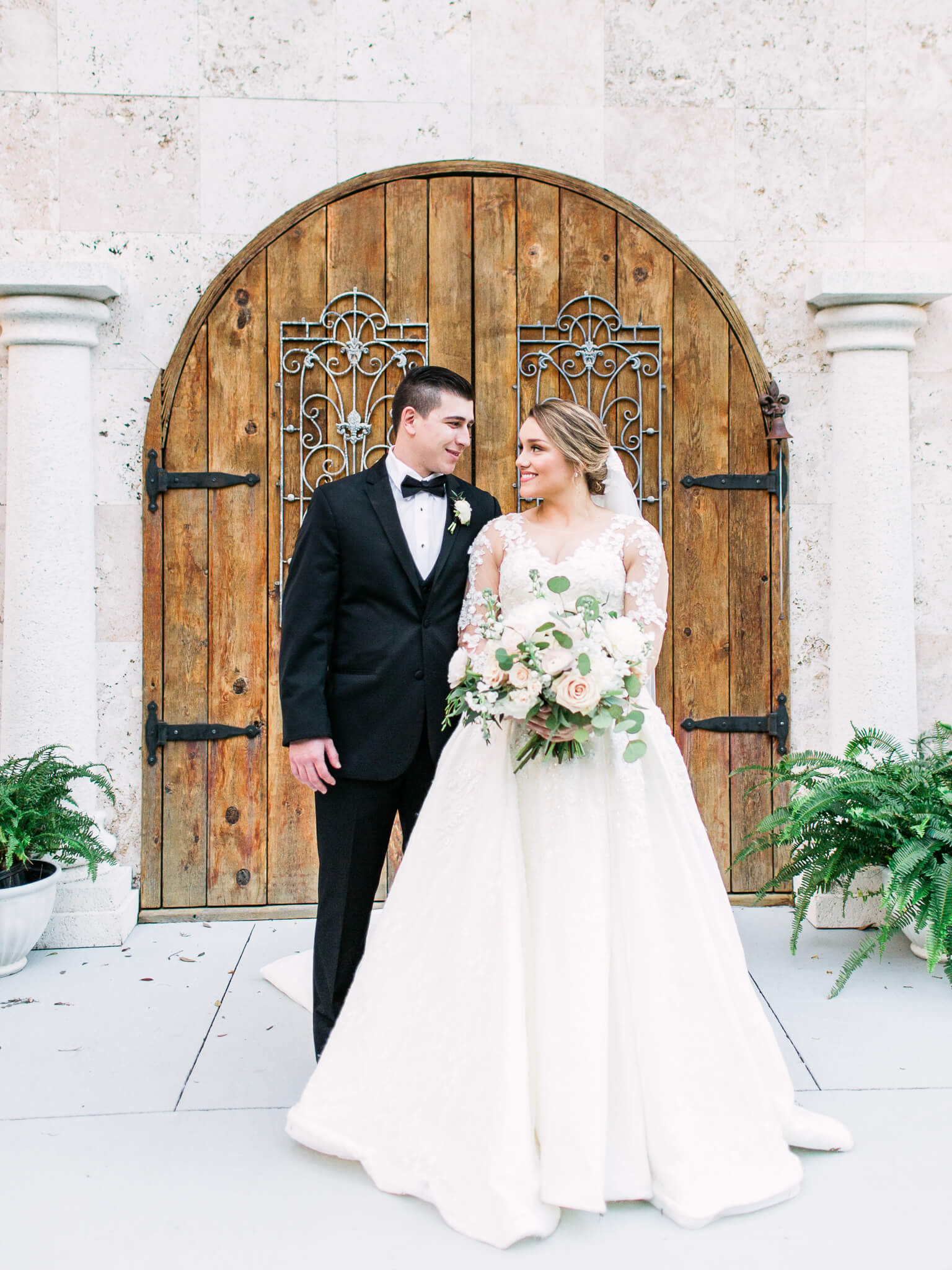 Groom and bride stand close together in front of rustic wooden doors while looking into each other with smiles 