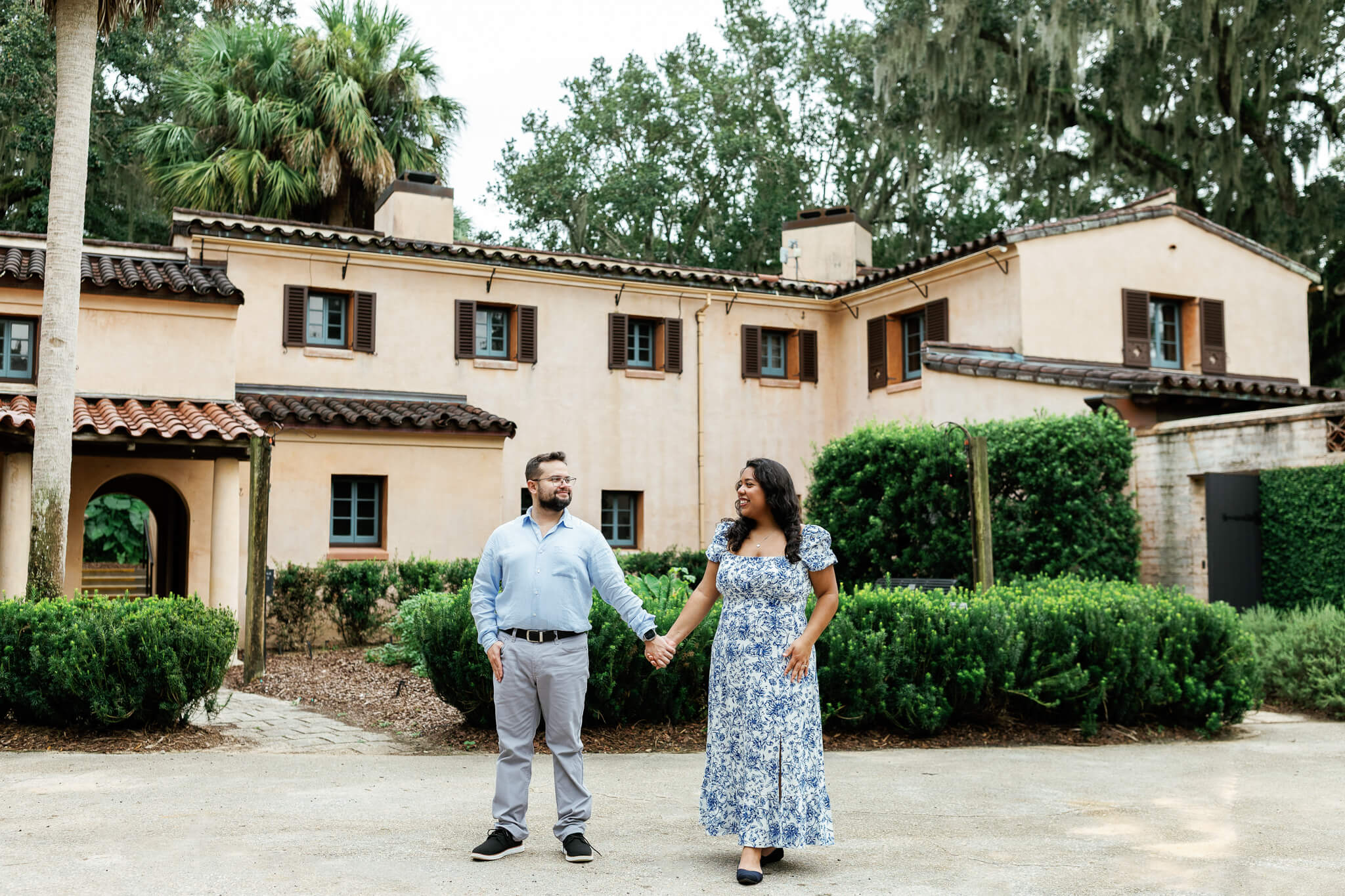 Couple holding hands in front of a mansion with green hedges