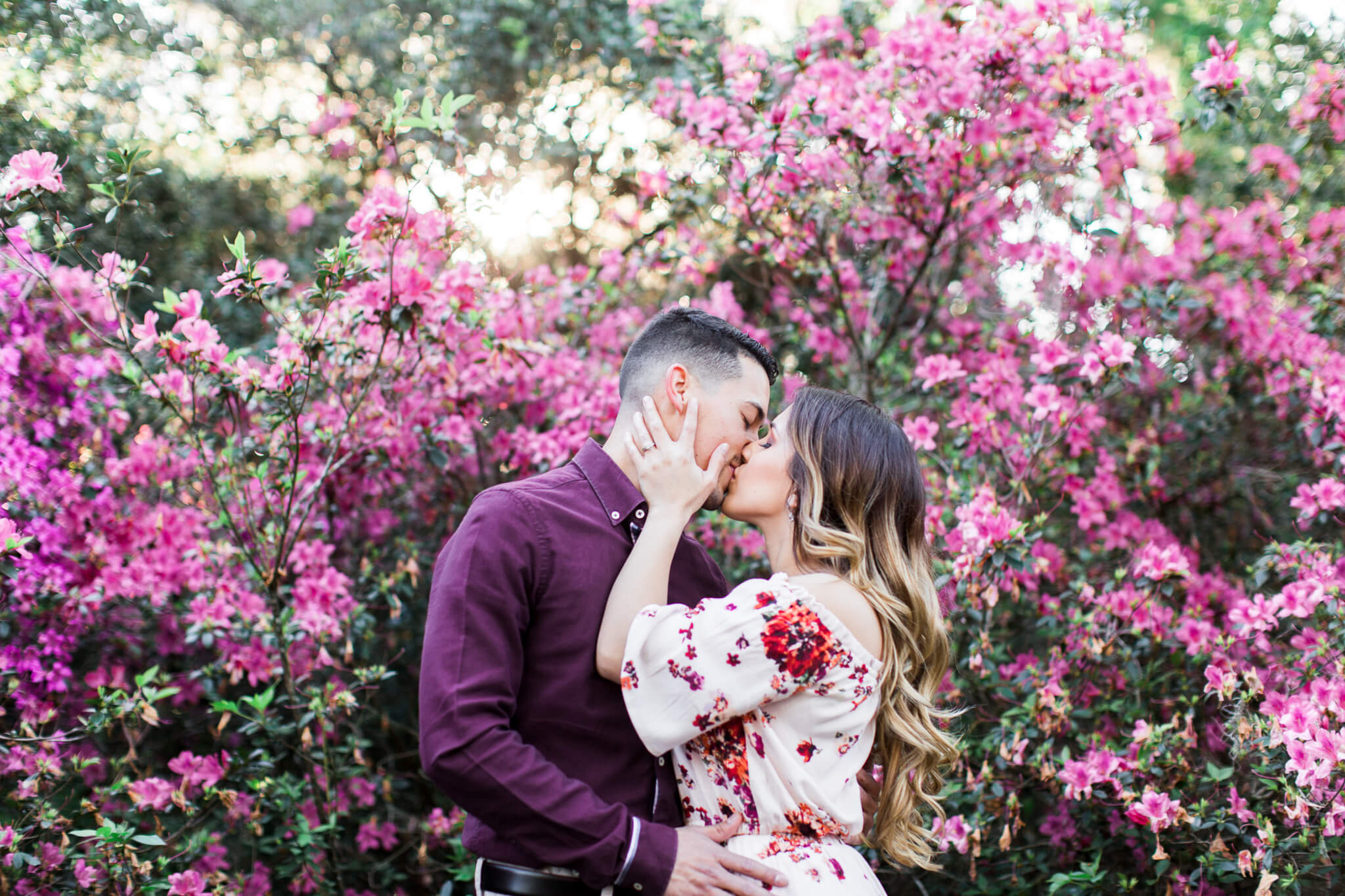 Engaged couple embracing and kissing in front of a large bloom of pink flowers during their engagement photos at Bok Tower Gardens