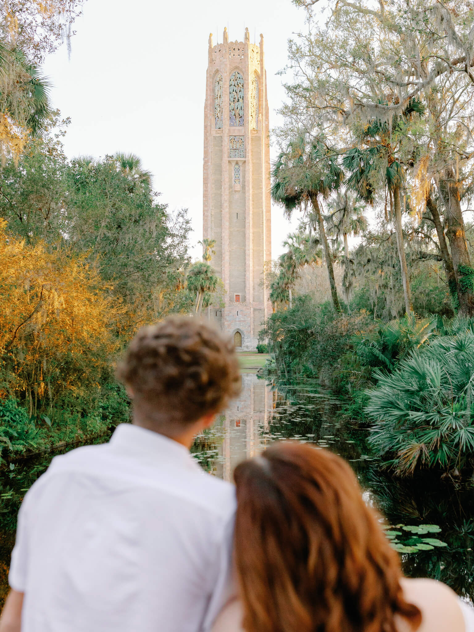 A couple standing close to each other look beyond a pond at a tower during their engagement session at Bok Tower