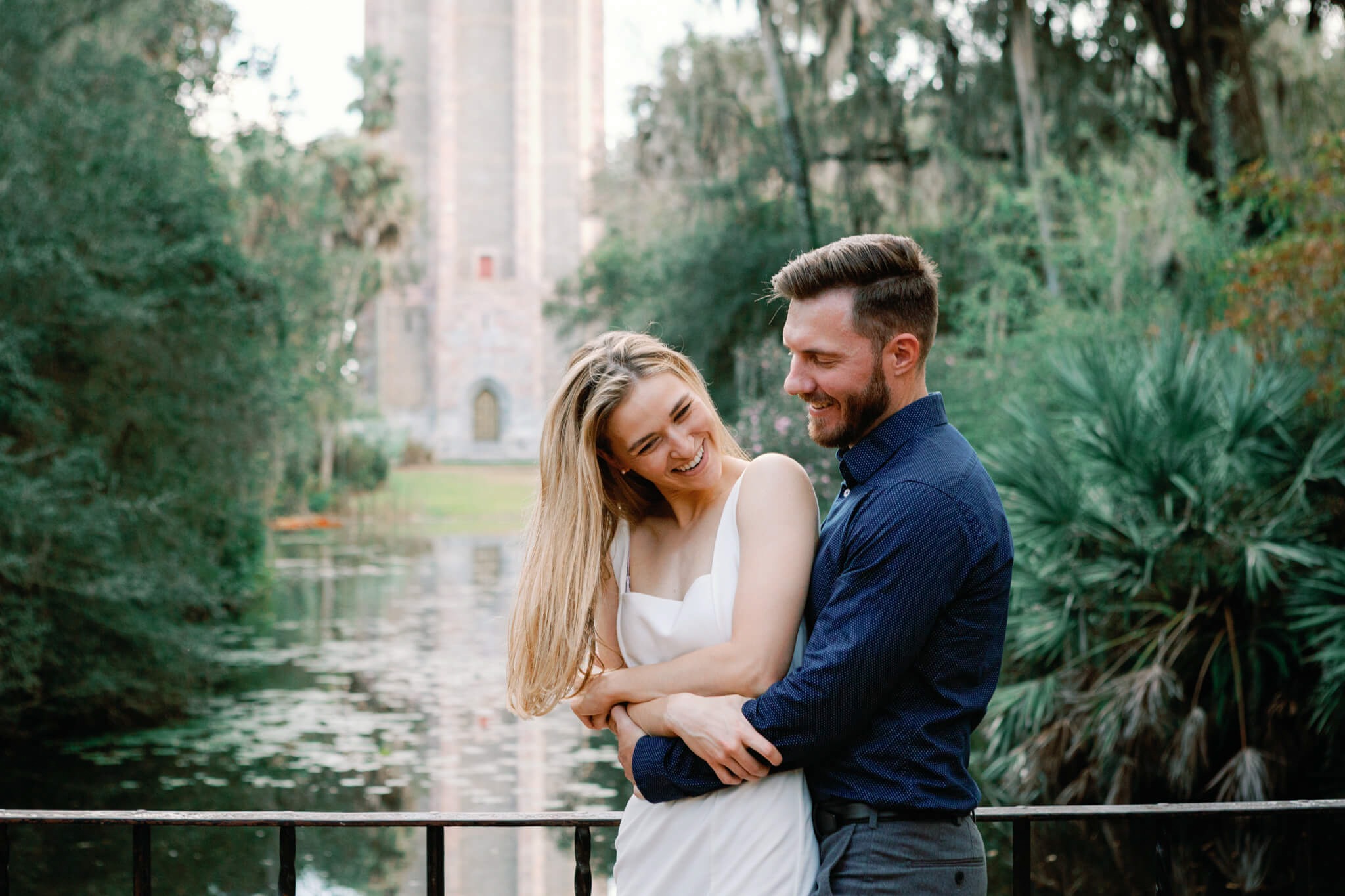 Engaged woman giggles as her boyfriend wraps her arms around her in front of a pond with a tower in the background during their engagement session at Bok Tower Gardens