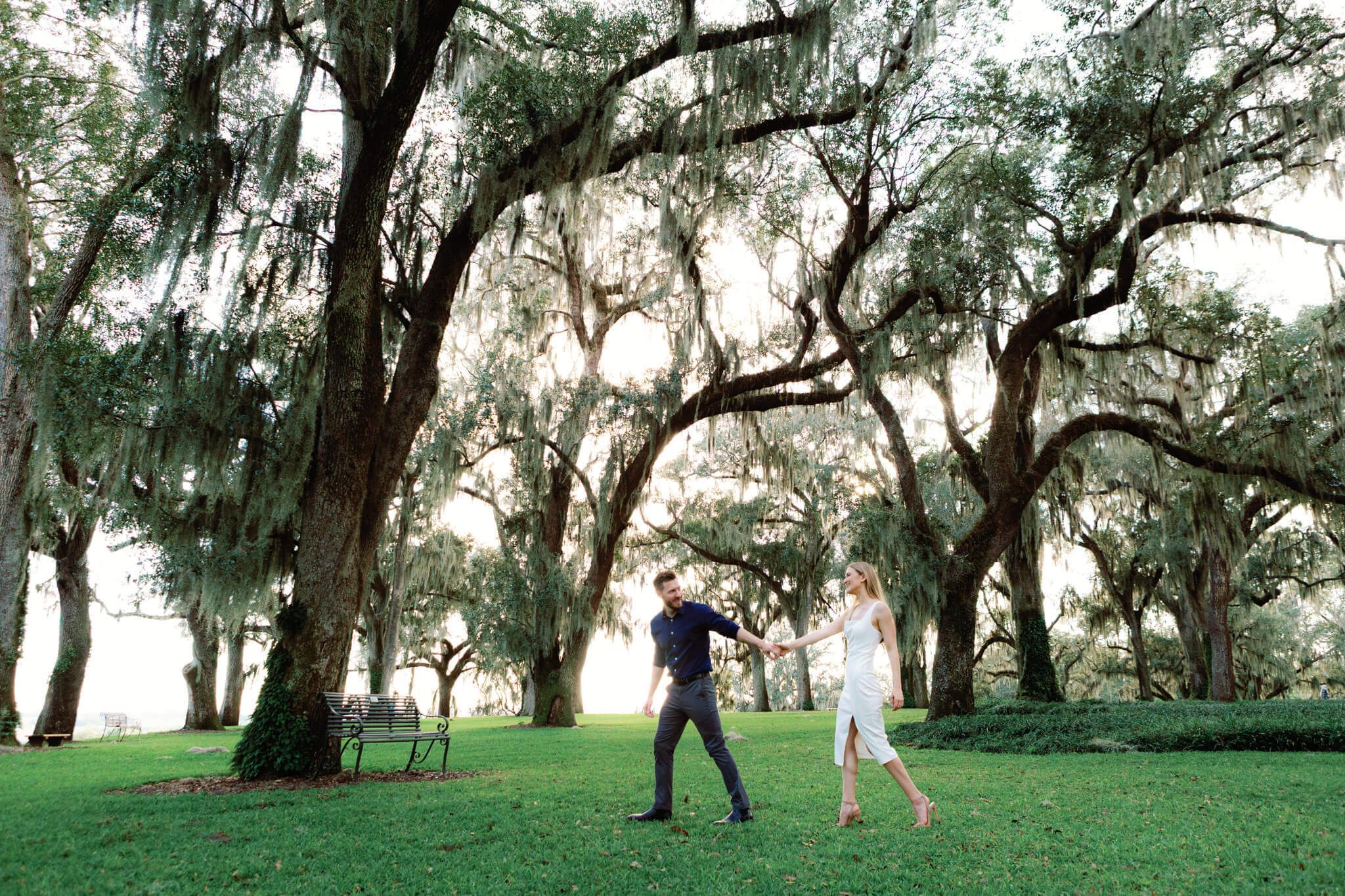 Man holds hand and leads girlfriend through a green lawn with tall trees during engagement session at Bok Tower Gardens