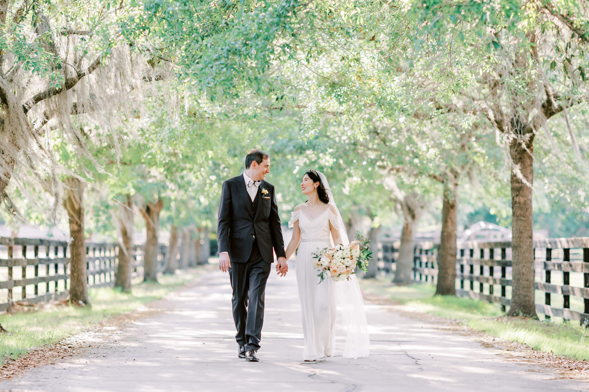 Bride and groom hold hands and walk on beautiful driveway surrounded by symmetrical trees on each side