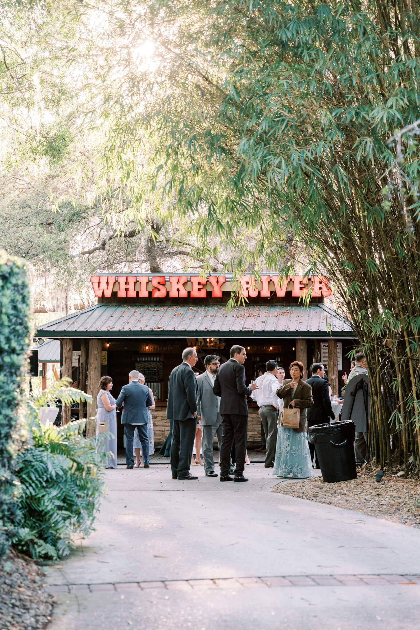 Wedding guests gather for cocktail hour in front of an outdoor bar that reads Whiskey River at an outdoor wedding venue in Central FL