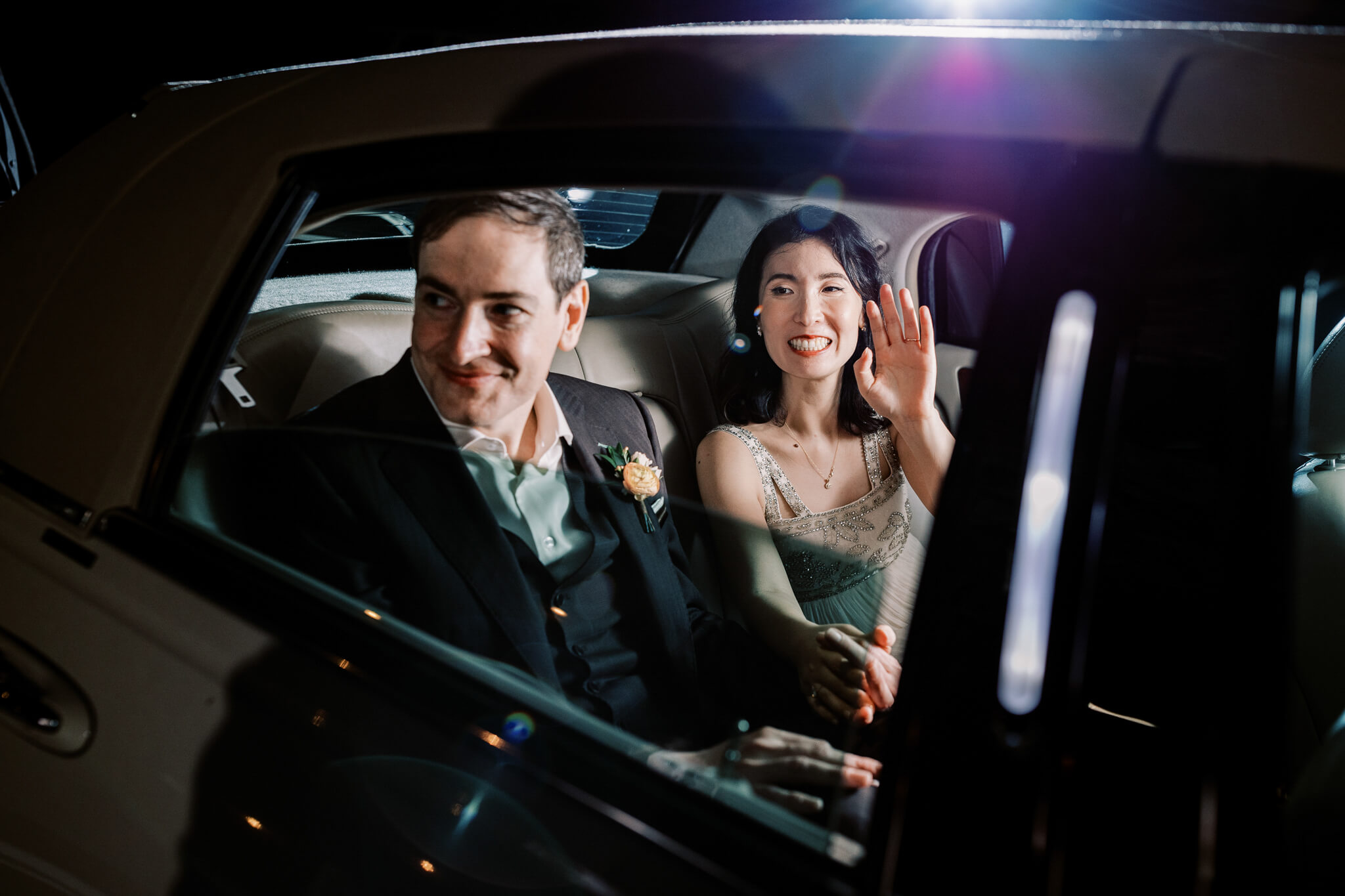 Bride and groom smile and wave goodbye to the wedding guests as they leave in a luxury car with the window halfway open