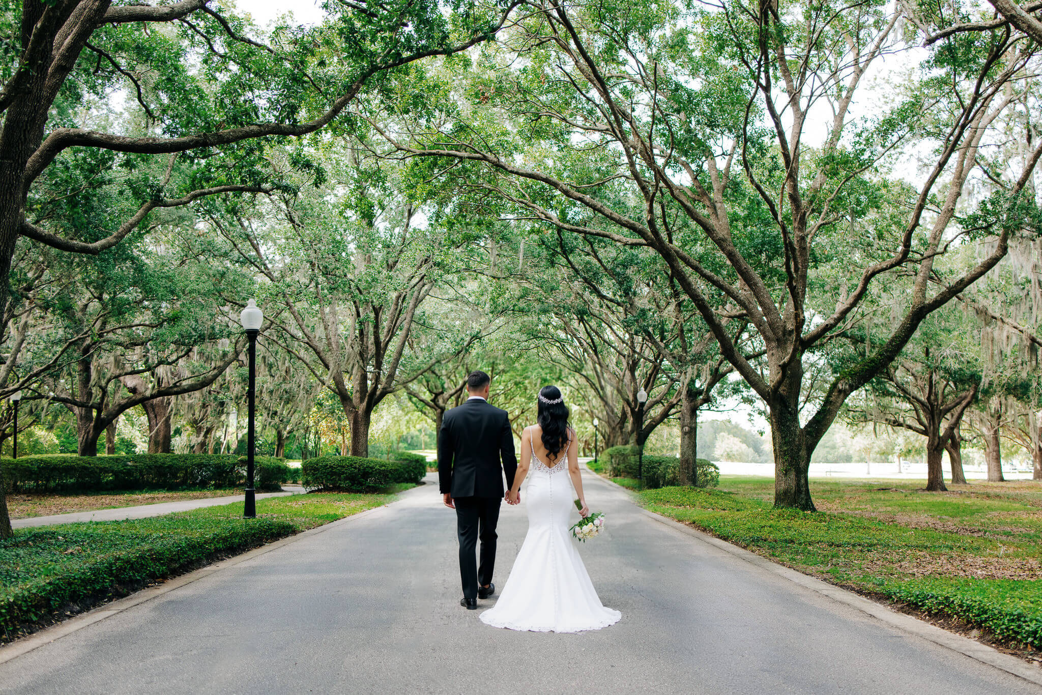 Groom holding bride's hand on a road surrounded by a lush canopy of trees at an Orlando wedding venue called Cypress Grove Estate House