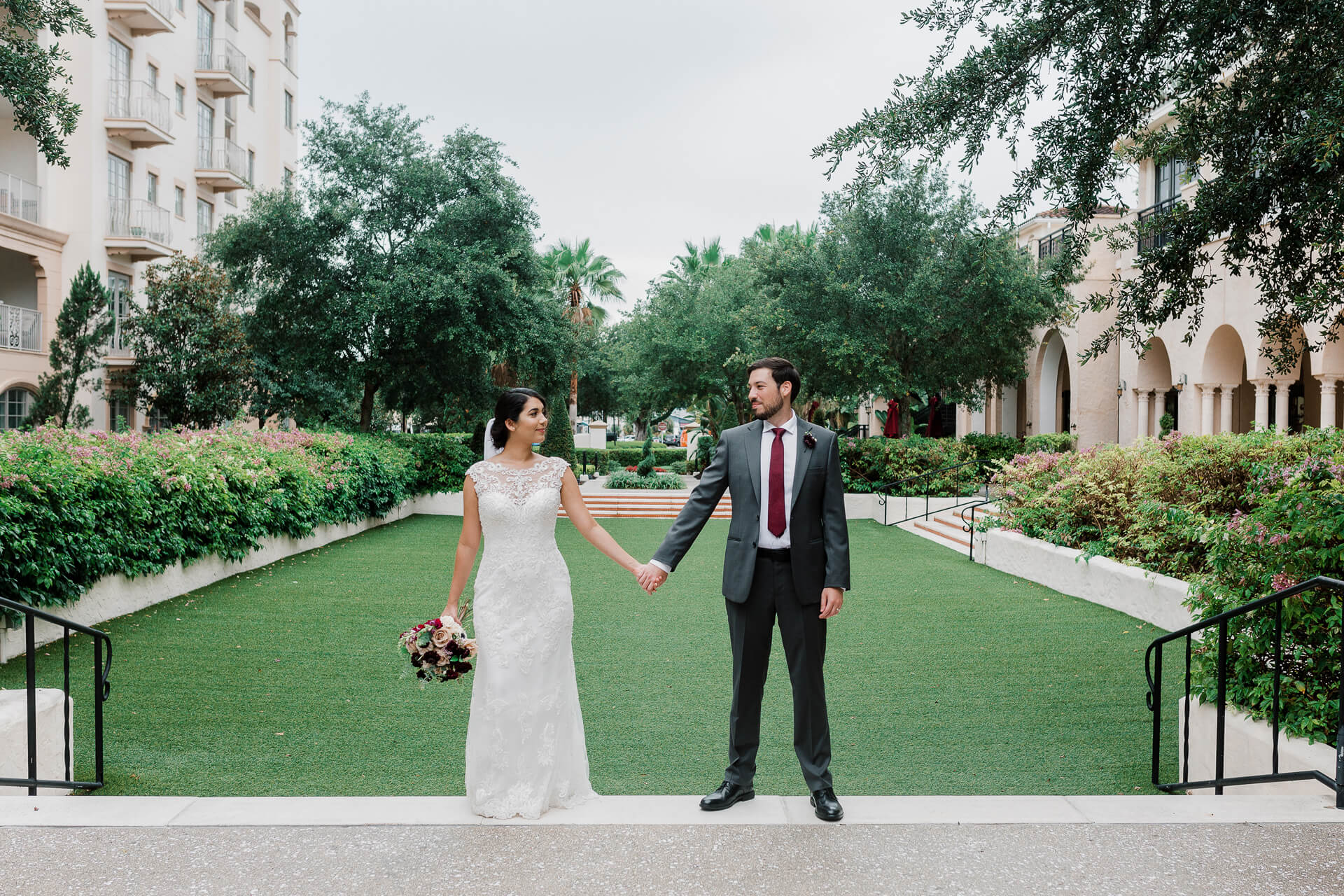 Bride and groom stand sideways and hold hands in an elegant wedding venue with a green lawn