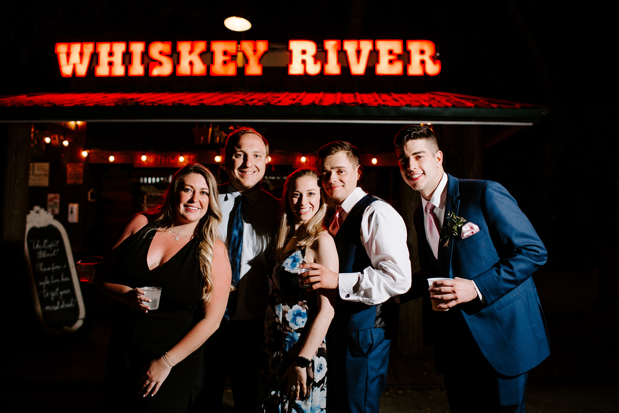 Wedding guests posing for a group photo in front of bar that with Whiskey River neon sign at Club Lake Venue