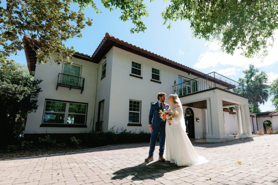 groom in blue suit next to bride in white wedding dress in front of a estate mansion on a sunny day at a wedding at Lake Lucy Estate