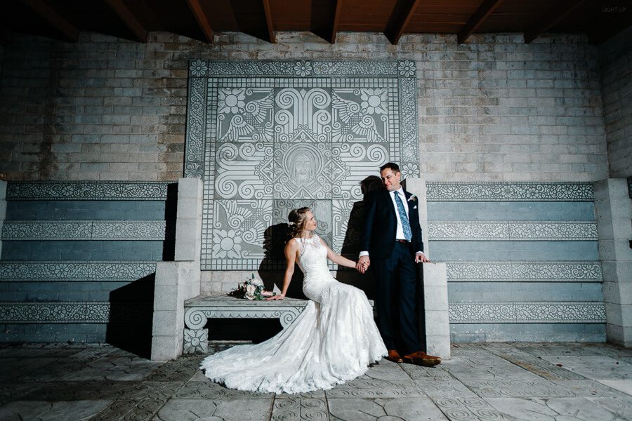 bride and groom pose for wedding photo in front of a wall with mayan textures and architecture